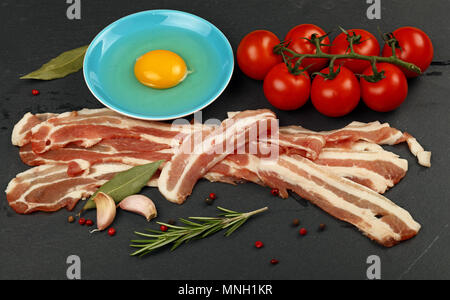 Les tranches de bacon de porc cru, tranches, d'œuf en bleu soucoupe, épices, poivre et tomates cerises fraîches rouge sur l'ardoise noire, Close up, high angle Banque D'Images