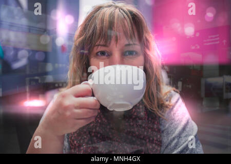 Une femme âgée de 40 ans de boissons dans une grande tasse de café. Elle se trouve derrière une vitre. Dans le reflet de la fenêtre, vous pouvez voir une gare. Banque D'Images