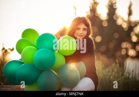 Portrait of a cute little girl sitting in the park avec des tas de ballons à air vert à l'extérieur dans une légère lumière soleil du soir, fêter anniversaire, preparati Banque D'Images