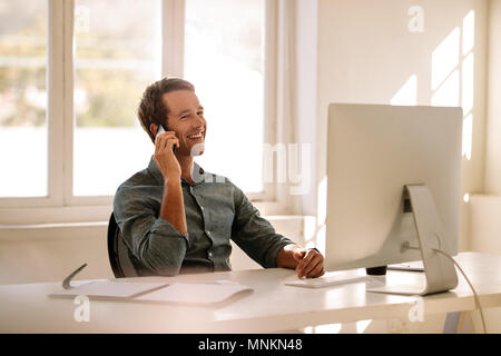 Smiling businessman talking on mobile phone tout en travaillant sur ordinateur. L'homme assis à sa table de travail travaillant sur ordinateur à la maison. Banque D'Images