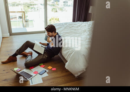 homme d'affaires lisant son journal tout en travaillant sur un ordinateur portable et en buvant du café. Homme assis sur le sol regardant son journal avec son ordinateur portable et offi Banque D'Images