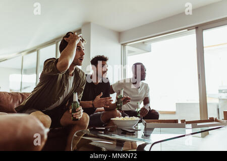 Trois amis de regarder un match à la télévision maintenant les bouteilles de bière. Les hommes assis sur un canapé avec des boissons et des collations en regardant la télévision à la maison. Banque D'Images