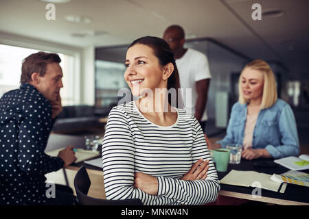 Young businesswoman looking par dessus son épaule et sourit alors qu'il était assis avec des collègues à une table dans un bureau Banque D'Images