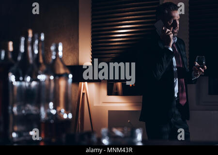 A souligné l'homme d'âge moyen d'un costume à parler au téléphone et la tenue d'un verre d'alcool tout en se tenant dans une pièce sombre avec des bouteilles d'alcool vides dans un lecteur blu Banque D'Images