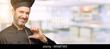 Cuisinier en chef, l'homme portant hat fier, heureux et fier, en pointant avec la victoire face à une cuisine de restaurant Banque D'Images