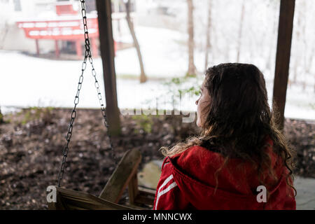 Closeup of young woman sitting on swing sous terrasse en bois de chambre sur cour dans quartier avec la neige durant tempête blanc blizzard, flocons Sächsischer Hof Banque D'Images