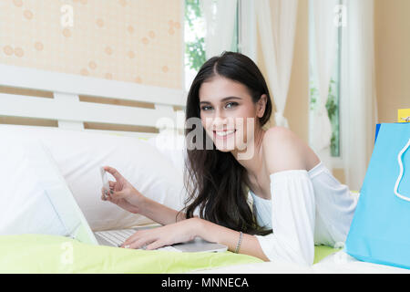 Photo of Asian woman doing online shopping sur ordinateur portable avec carte de crédit au lit à la maison. Banque D'Images