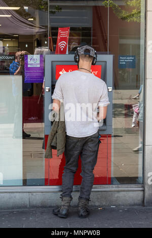 Un jeune homme portant une casquette de baseball et un casque à l'aide d'une machine à cahpoint ou un trou dans le mur de guichet automatique bancaire distribanque. Banque D'Images