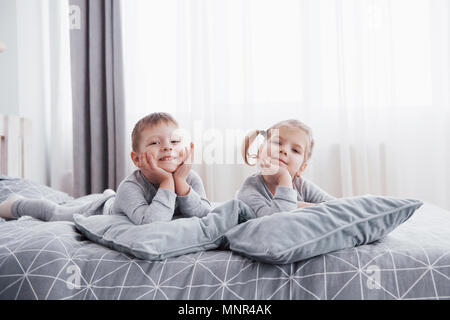 Heureux les enfants jouant dans la chambre blanche. Petit garçon et fille, frère et sœur jouer sur le lit wearing pajamas. L'intérieur de pépinière pour les enfants. Vêtements et literie pour bébé et enfant. Famille à la maison Banque D'Images