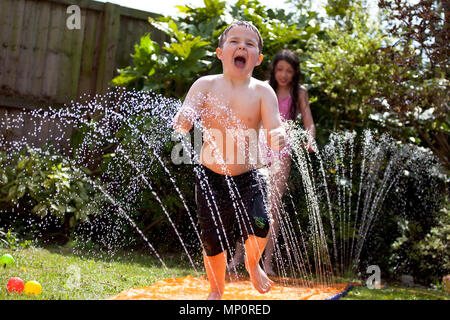 Jeune garçon jouant avec aspersion d'eau dans le jardin arrière Banque D'Images