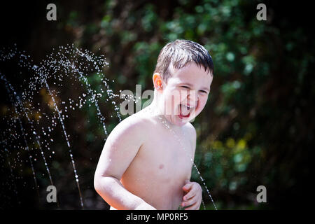 Jeune garçon jouant avec aspersion d'eau dans le jardin arrière Banque D'Images