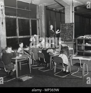 Février 1949 pictiure historique, d'un junior d'après-guerre, de classe d'école avec enseignante à un tableau noir, assis à un bureau d'enseignement childern, à l'intérieur d'une ancienne usine avec de grandes fenêtres en métal, Essex, Angleterre, Royaume-Uni. Banque D'Images