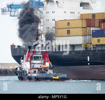 Tug boat éructations de fumée noire comme il guide SMC Floriana porte-conteneurs au port de Las Palmas de Gran Canaria, Espagne. Banque D'Images