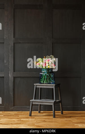 Un riche bouquet coloré de belles fleurs d'été dans un vase de verre bleu sur un escabeau noir dans une sombre salle de séjour intérieur Banque D'Images