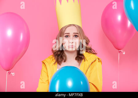 Jeune femme déçue dans party hat entouré de ballons colorés, isolé sur fond rose pastel. Triste Anniversaire concept. Banque D'Images