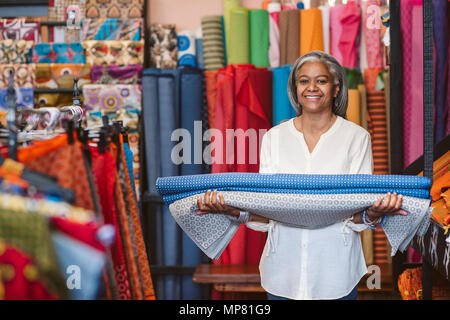 Smiling mature woman holding rouleaux de tissu dans son magasin de tissus Banque D'Images