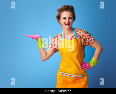 Grand temps de nettoyage. souriante jeune femme au foyer dans un tablier jaune présentant quelque chose sur palm vide isolé sur fond bleu Banque D'Images