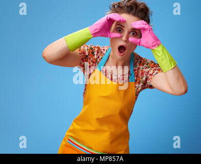 Grand temps de nettoyage. surpris femme moderne dans un tablier jaune à la main en forme de jumelles par isolé sur blue Banque D'Images