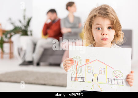 Bouleversé petit garçon tenant un dessin d'une maison, avec ses parents en colère assis sur un canapé dans l'arrière-plan flou Banque D'Images