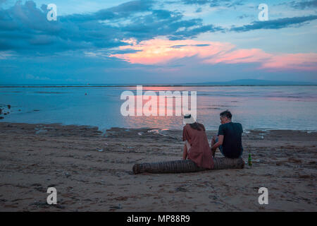 Une femme et un homme assis sur un tronc d'arbre sur la plage au coucher du soleil, une bouteille de bière se distingue par le côté, Sanur, Bali, Indonésie, Apr Banque D'Images