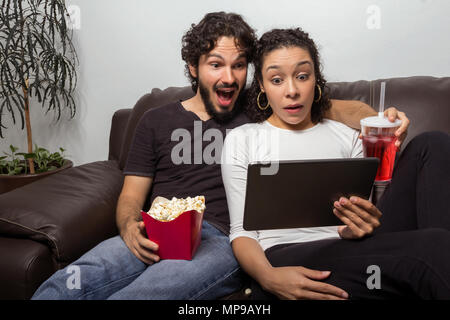 Effrayé et surpris couple est regarder film streaming sur tablette. Eating popcorn et boisson. Ils sont assis sur le canapé à la maison. Lieu de loisirs à Banque D'Images