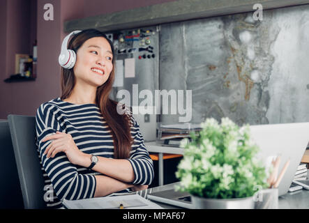 Young Asian businesswoman décontractée sur le bras de bureau reste posent avec ordinateur portable et l'écoute de la musique via la prise casque et de regarder à travers la fenêtre de bureau ,travailler Banque D'Images