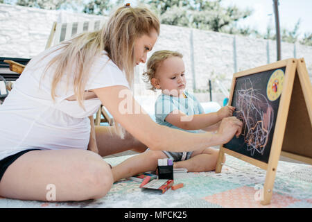 Le portrait d'une femme enceinte bénéficiant d'griffonner avec son fils sur un tableau. Une mère d'aider son bébé garçon en s'appuyant sur un tableau. Banque D'Images