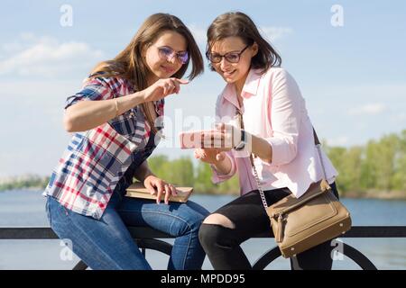 Deux heureux female friends watching photos, vidéos sur le smartphone et avoir du plaisir. Fond nature, parc, rivière. Mode de vie urbain et l'amitié conc Banque D'Images