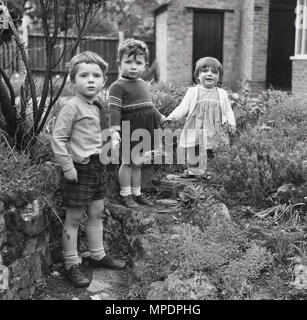 1961, historiques, trois enfants tenir la main tout en jouant dans la rocaille d'un jardin à l'arrière d'une grande maison à Hanwell, Ealing, à l'ouest de Londres, en Angleterre. UK. Banque D'Images