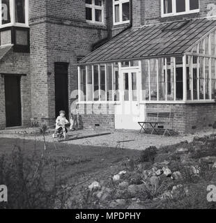 1961, historique, petit garçon monte un tricylce sur une zone de gravier dans le jardin à l'arrière d'une grande maison à Hanwell, Ealing, à l'ouest de Londres, en Angleterre. UK. Banque D'Images