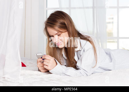 Jeune fille se trouve dans une chemise d'homme sur le lit avec un smartphone dans les mains de Banque D'Images