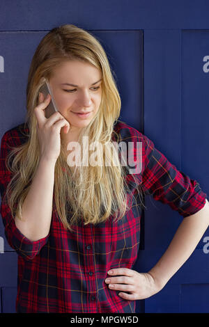 Jolie femme appeler quelqu'un par téléphone portable en souriant à la caméra contre mur bleu Banque D'Images