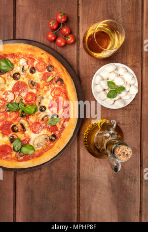 Photo prise à la verticale de la pizza au pepperoni, avec verre de vin blanc, huile d'olive, tomates cerises et mozzarellini fromage, tourné à partir de ci-dessus sur dark background rustique Banque D'Images