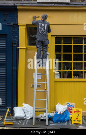 Un homme à l'aide d'une échelle pour peindre un bâtiment jaune. L'homme sur une échelle sur le côté d'un bâtiment peintre et décorateur au travail la peinture des couleurs vives. Banque D'Images