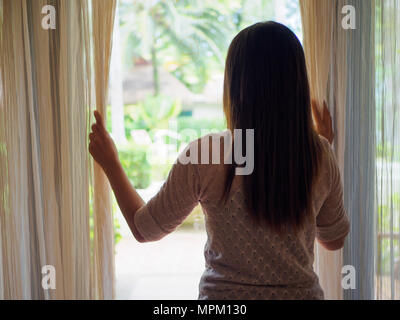 Vue arrière d'une jeune femme tenant les rideaux ouverts à regarder dehors d'une grande fenêtre d'éclairage à la maison, de l'intérieur. Ambitieux et positif de vie. Femme Banque D'Images