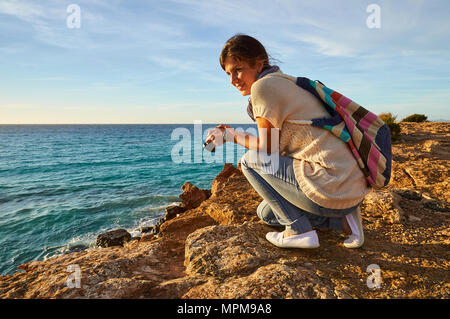 Jeune femme avec un appareil photo dans ses mains et genoux, observant la mer au coucher du soleil près de Cala Saona à Formentera (Baléares, Espagne) Banque D'Images