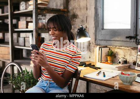 Souriante jeune femme assise en face de bureau dans un loft looking at cell phone Banque D'Images