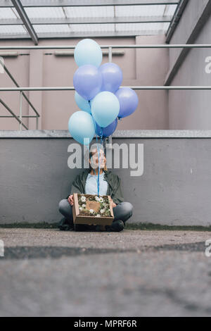 Femme souriante avec des fleurs en boîte en carton et bouquet de ballons bleu assis sur sol Banque D'Images