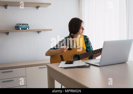 Young woman sitting at table at home with laptop playing guitar Banque D'Images