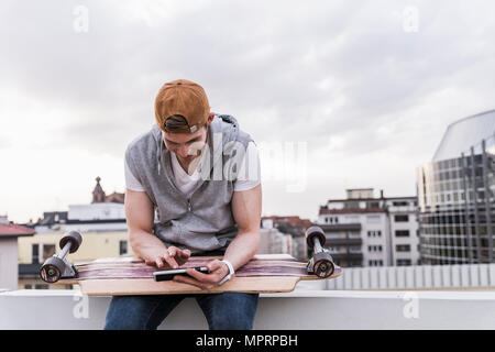 Smiling man sitting dans la ville avec skateboard using cell phone Banque D'Images