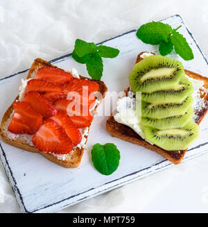 Deux toasts avec du caillé doux, tranches de fraise et kiwi sur une planche de bois blanc, vue du dessus Banque D'Images