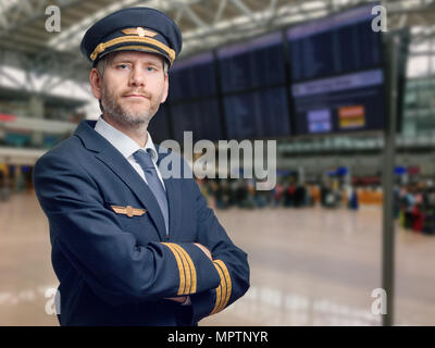Portrait d'un pilote en uniforme avec des rayures d'or et cap croisa les bras. Il est debout dans le terminal d'un aéroport Banque D'Images