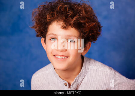 Close-up portrait of smiling preteen boy avec les cheveux bouclés et les yeux bruns contre fond bleu Banque D'Images