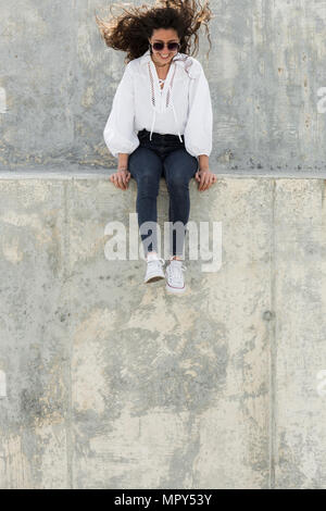 Toute la longueur de happy woman wearing sunglasses, assis sur mur de retenue Banque D'Images