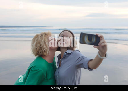 Cheerful woman with smart phone selfies lors d'être embrassé par ami à la plage pendant le coucher du soleil Banque D'Images