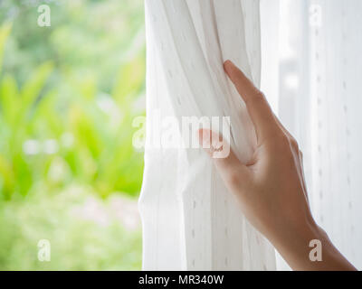 Woman's hand ouvrant les rideaux dans la chambre avec la lumière naturelle et le jardin arrière-plan. Banque D'Images