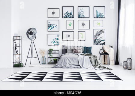 Photo réelle d'un lit debout entre une lampe et une chaise dans une chambre lumineuse intérieur avec tapis noir et blanc et des affiches sur un mur Banque D'Images