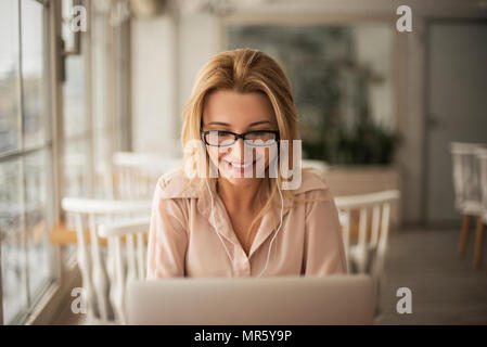 Jeune femme blonde portant des lunettes et d'écouteurs en café assis à écouter de la musique de table working on laptop smiling smiling close-up bl Banque D'Images