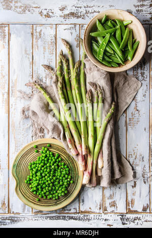 Variété de matières organiques non cuit les jeunes légumes verts asperges, petits pois, pois pod dans des assiettes en céramique sur lin tissu sur planche en bois blanc arrière-plan. T Banque D'Images