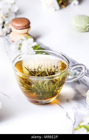 Tasse en verre de thé vert chaud avec dessert français macarons, fleurs de printemps magnolia blanc et branches en fleurs de cerisier sur le marbre blanc de la texture background Banque D'Images